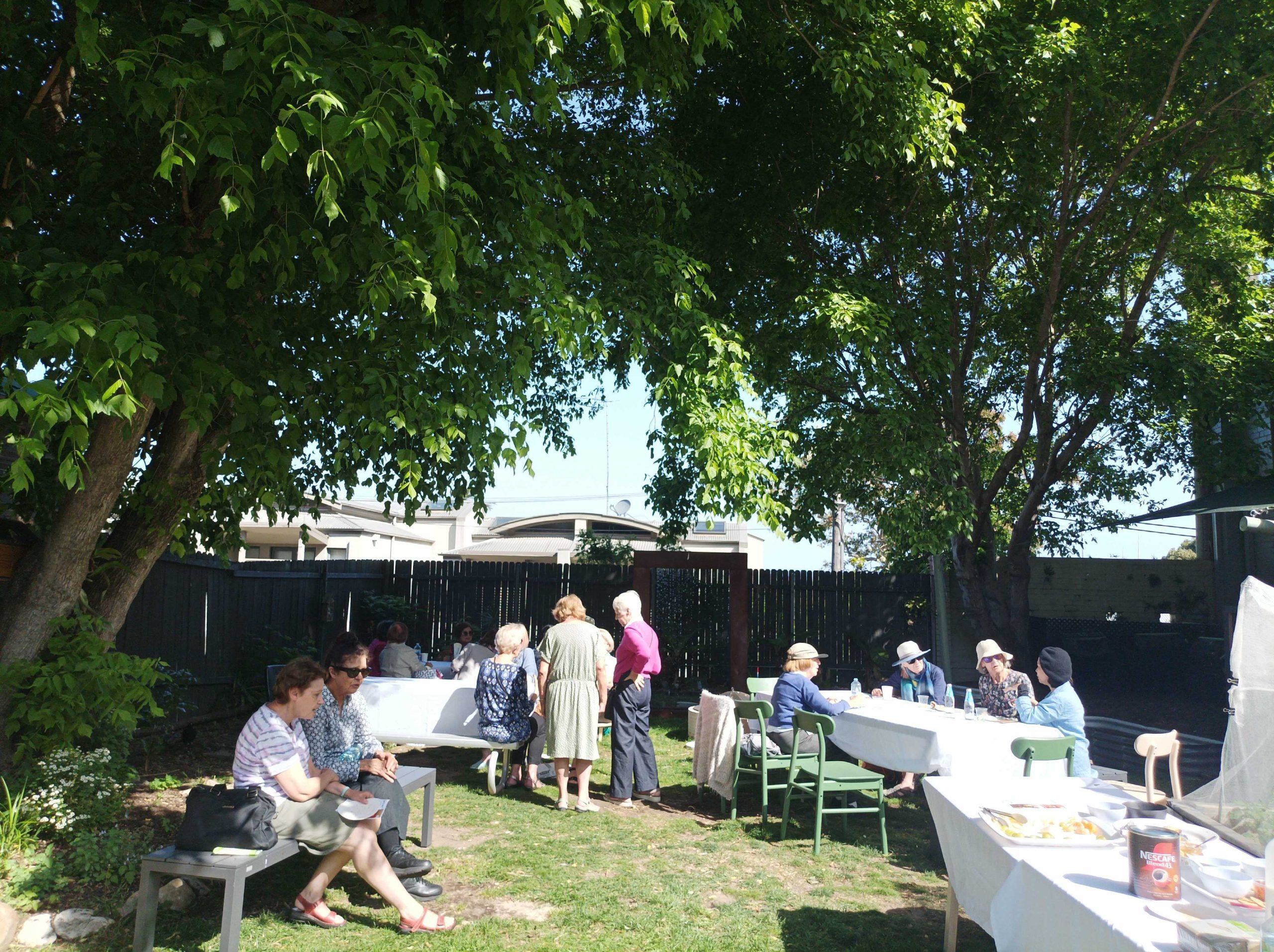 People sitting and talking at long tables set out under trees in a sunny garden