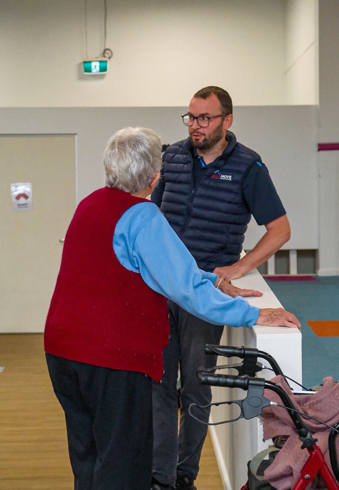 A worker talking with an older woman who is steadying herself on a walking frame in a community hall