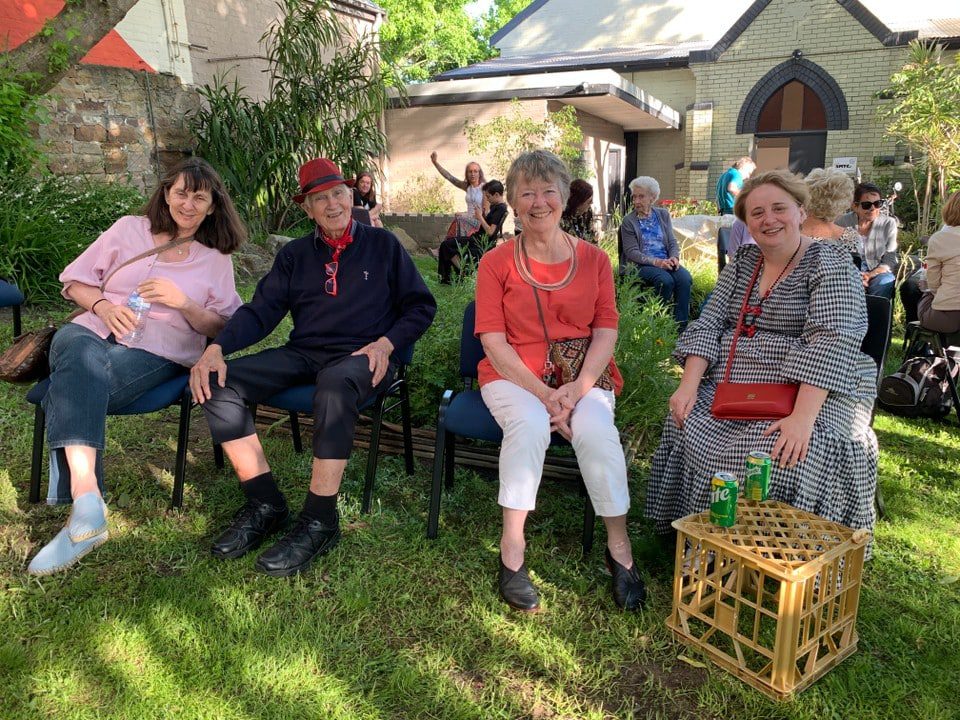 Four people sitting on chairs on the grass at an outdoor community event, bunting strung overhead