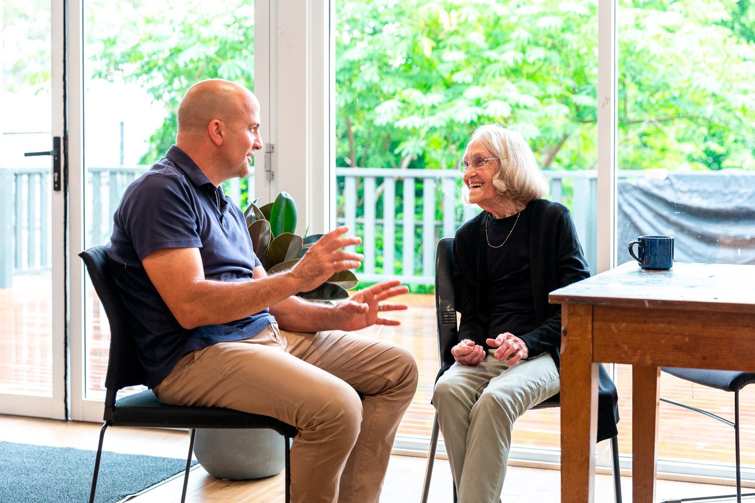 A man and an older woman talking, seated by a window