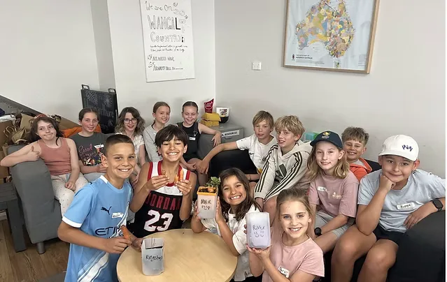 A group of children sitting together around a low table, smiling and holding decorated jars they have made. An Acknowledgement of Country poster and a map of Aboriginal Australia hang on the wall behind them.