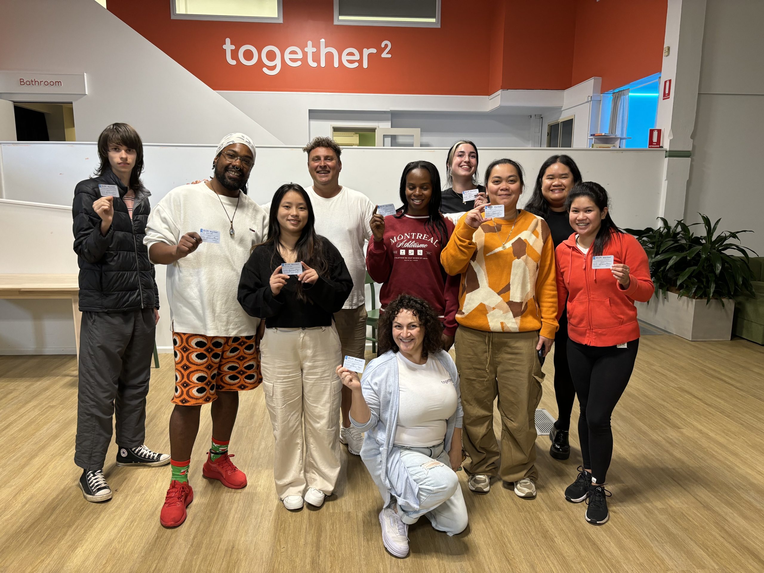 Ten together² team members standing together in front of the together² wall at the Rozelle hub, each holding up a card