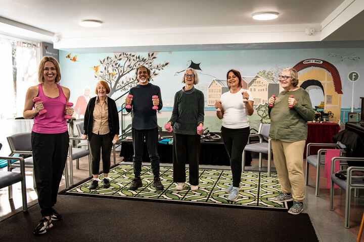 Six people standing in a circle with hand weights during an exercise group in a community room