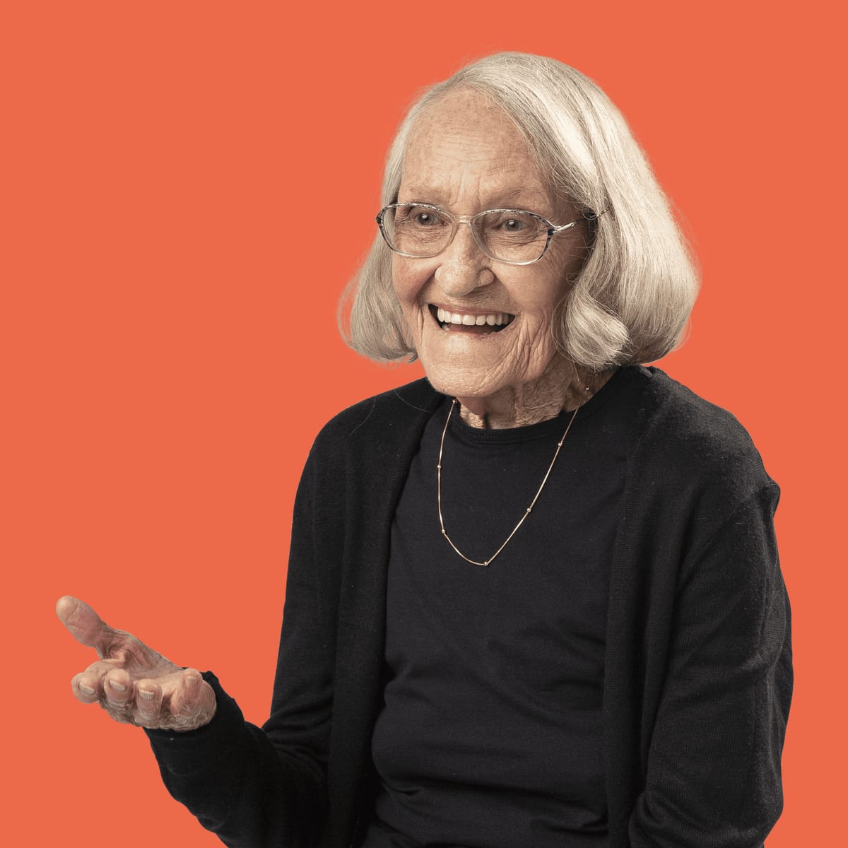 Portrait of an older woman with white hair and glasses, smiling warmly with one hand open in front of her